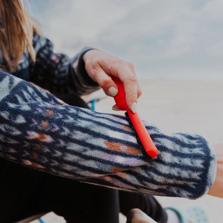 Using the Lilly Brush - Be Forever Furless Mini, a person refreshes winter clothes by cleaning the sleeve of a patterned fleece jacket. The scene is set outdoors with sandy ground and a blurred sky in the background.