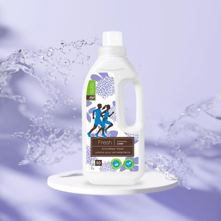 A bottle of Fresh® Activewear Wash by Fashion Care sits on a circular platform surrounded by water splashes. The label highlights workout gear and odor protection with running figures, fashion care emphasis, fluid ounces, and eco-friendly symbols, underscoring its 1L (33.8 oz) eco-friendly ingredients.