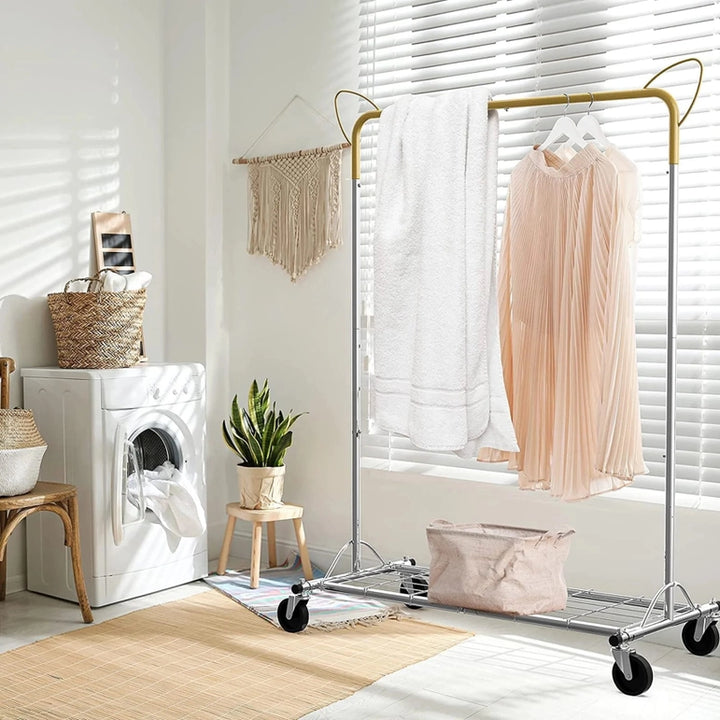 A bright laundry room showcases the Above Edge Hanging Garment Rack with Shelf, holding a pink dress and white towel. A woven basket and potted plant sit nearby. Sunlight filters through window blinds, casting shadows on the floor, while a macramé wall hanging enhances the charm.