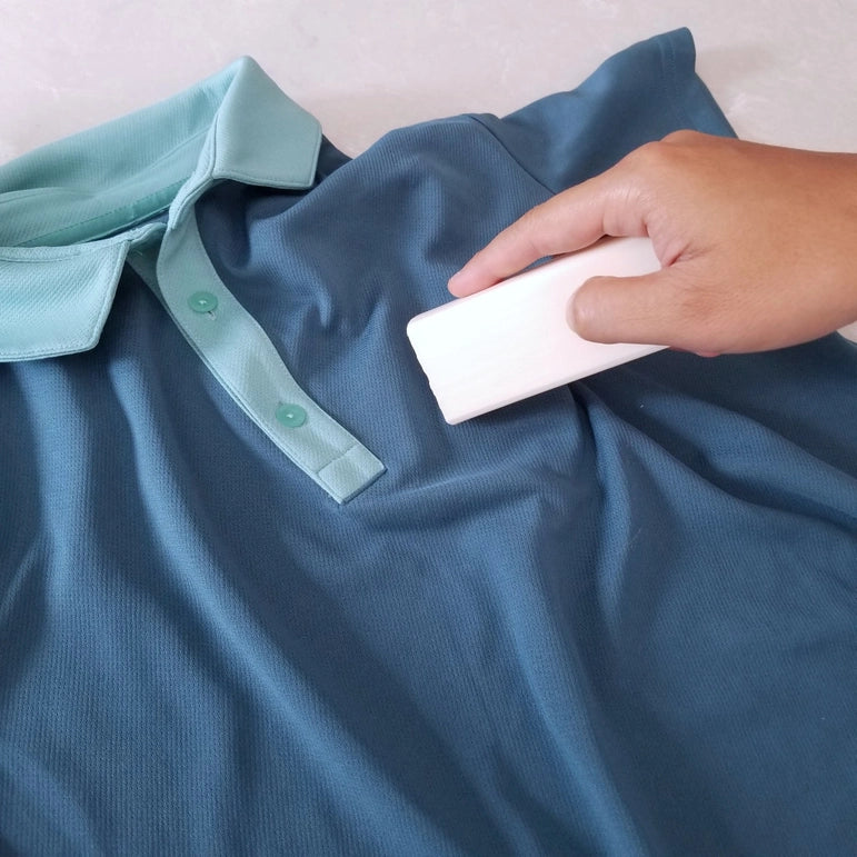 A hand uses a Vegan Laundry Stain Remover Bar by Kind Laundry on a folded blue polo shirt with a light teal collar.