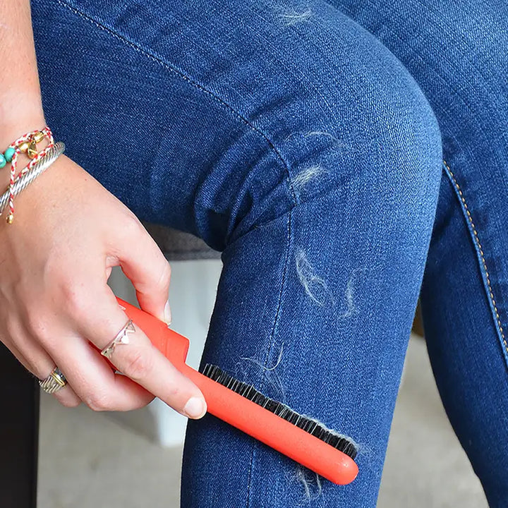 Adorned with a colorful bracelet and rings, a person deftly uses the red Lilly Brush - Be Forever Furless Mini by Lilly Brush to swiftly remove pet hair from blue jeans, demonstrating its effectiveness in cleaning the fabric.