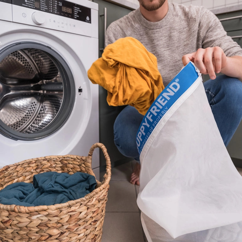 A person crouches near a washing machine with a yellow garment and the GUPPYFRIEND Washing Bag-Medium 19.7 in. x 29.1 in., designed to reduce fiber shedding and combat microplastics, while a wicker basket with blue clothes rests on the tiled floor among green cabinetry.