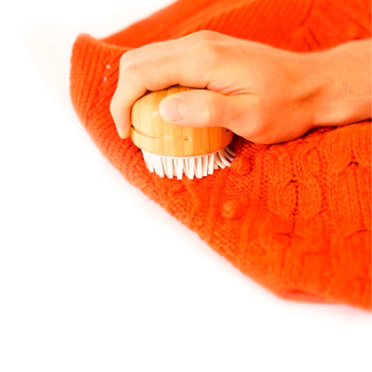 A hand uses the Fuzz Off Reusable Lint Remover by Full Circle Home with white bristles to gently clean a bright orange cable-patterned knitted fabric, efficiently removing pet hair as it glides smoothly against the white backdrop.