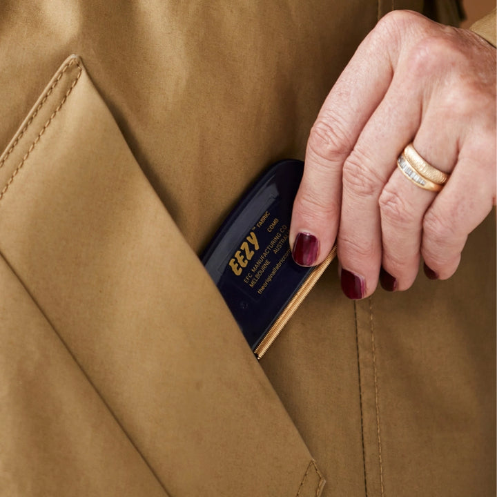 With red nails and a gold ring, someone slips a sleek black object labeled The Original Fabric Comb into the left pocket of a tan coat, ready to tackle fuzzy fabric easily with the innovative EZY™ Fabric Comb.