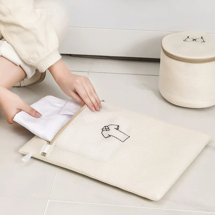 A person places a white cloth into an All Things Laundry beige mesh wash bag from the Set of 6 Mesh Laundry Wash Bags, designed to prolong garment life. The securely zipped bag has a shirt icon. Nearby, a round mesh laundry basket with a lid sits on light-colored tiles, ready to protect delicates.