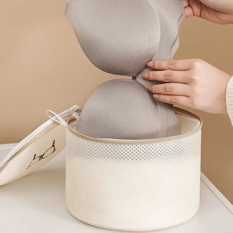 A person gently places a gray bra into the All Things Laundry mesh wash bag, part of the Set of 6, perfect for protecting delicates. This round container, with a black line drawing on its lid, extends the life of clothes.