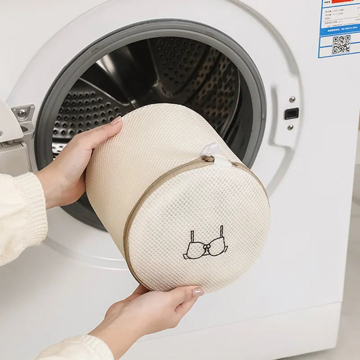 A person holds a beige cylindrical mesh laundry wash bag from All Things Laundrys Set of 6 in front of an open washing machine, featuring a delicate items icon. Ideal for protecting delicates like bras, this bag helps extend garment life while keeping them safe during washes.