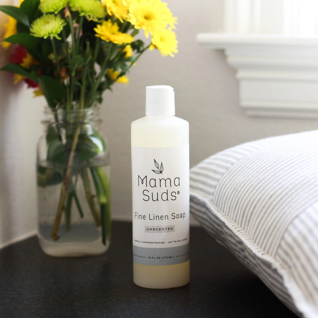 A 16 oz bottle of Mama Suds Fine Linen Soap-Unscented stands next to a striped pillow on a dark surface, with a vase of yellow and white flowers by a window in the background.