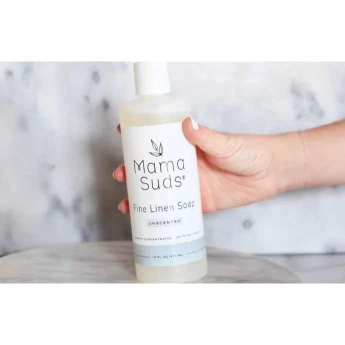 A hand holds Mama Suds Fine Linen Soap-Unscented, 16 oz, labeled as all-natural. Against a soft marble-like background, this non-toxic soap ensures gentle fabric care.