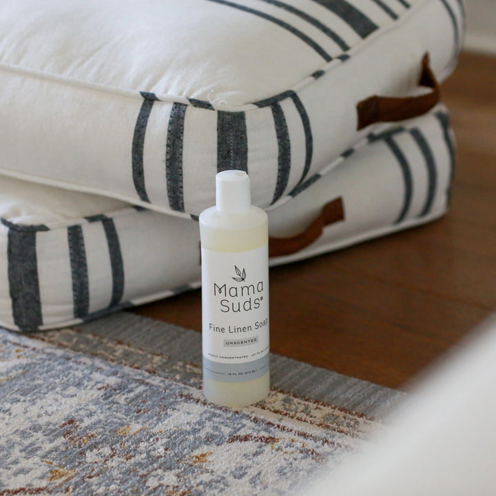 A 16 oz bottle of Mama Suds Fine Linen Soap-Unscented rests on a patterned carpet in front of stacked white and gray striped cushions with leather handles against a wooden floor.