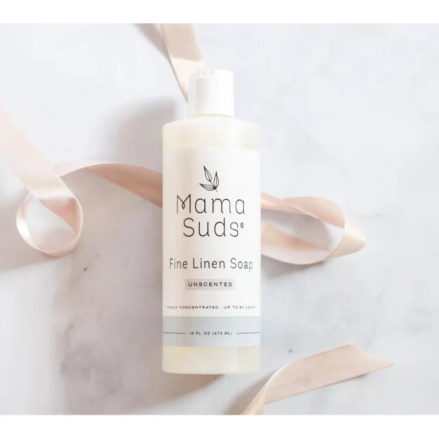 A 16 oz bottle of Mama Suds Fine Linen Soap-Unscented sits on a marble surface, adorned with a soft pink ribbon to emphasize its biodegradable and non-toxic nature.