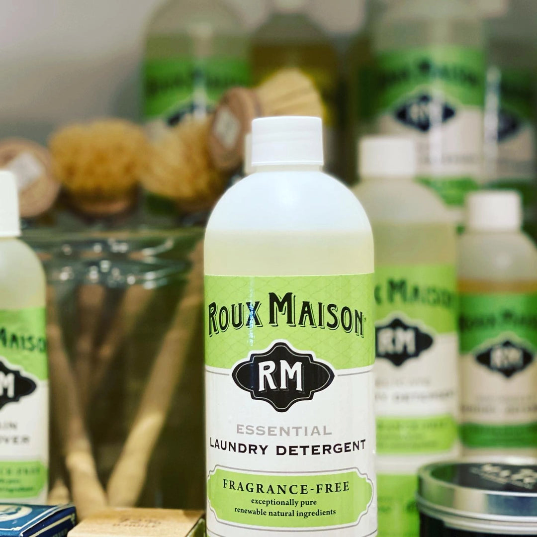 Close-up of Roux Maison Essential Laundry Detergent, fragrance-free and 1,4 dioxane free in a 16 oz bottle with a green and white label, surrounded by cleaning products and brushes. Made with natural ingredients and biodegradable for an eco-friendly clean.