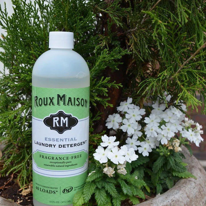 Amidst lush greenery and white flowers, a 16 oz bottle of Roux Maisons Essential Laundry Detergent stands out. Its marked for 40 loads and is fragrance-free, biodegradable, eco-friendly, all-natural, and free from 1,4 Dioxane.