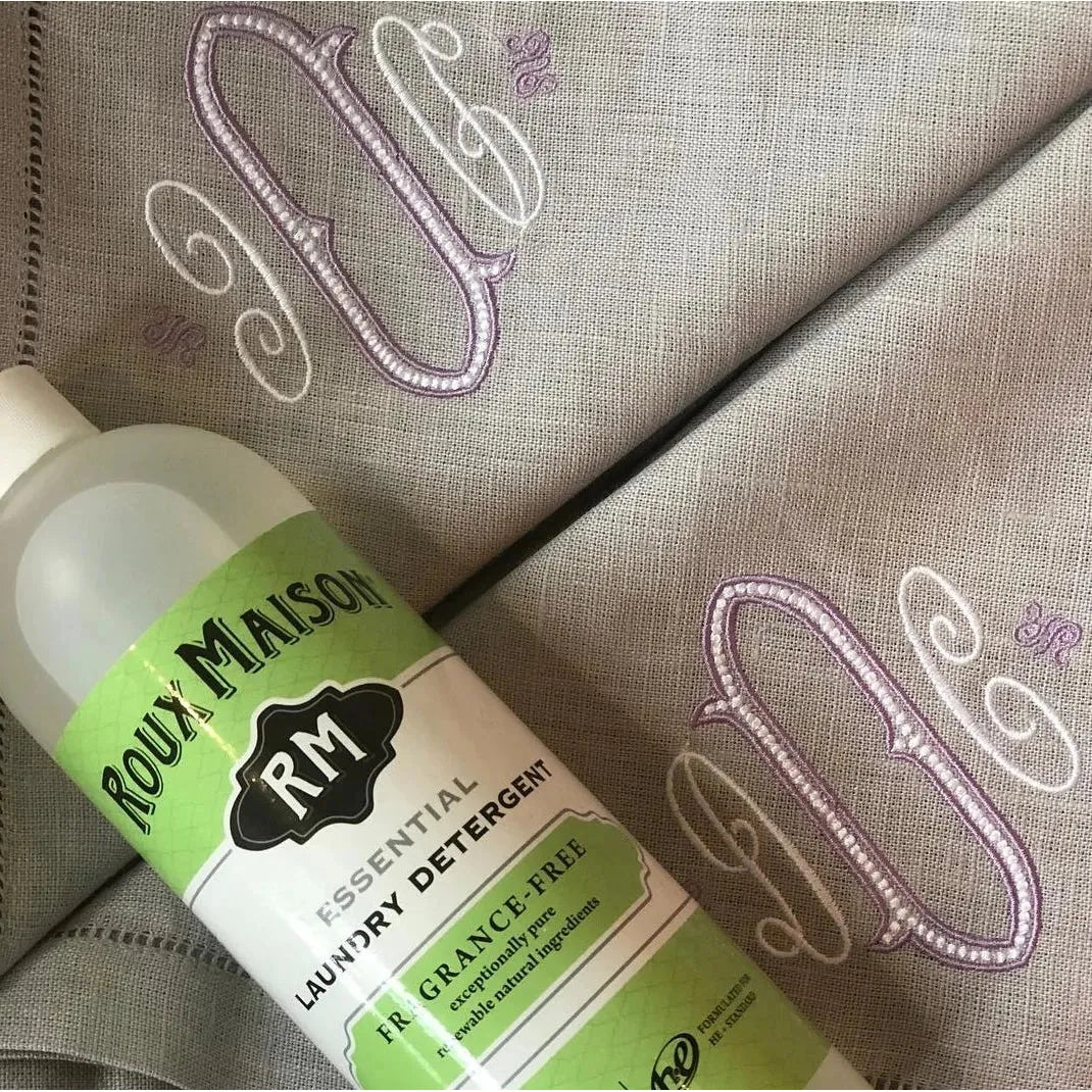 A 16 oz bottle of Essential Laundry Detergent by Roux Maison, fragrance-free and 1,4 Dioxane free, rests on gray linen with purple and white embroidery.