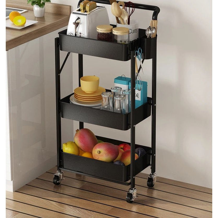 The black All Things Laundry 3 Tier Folding Cart holds kitchen items next to a counter: a toaster and utensils on top, plates and jars in the middle, and mangoes below.