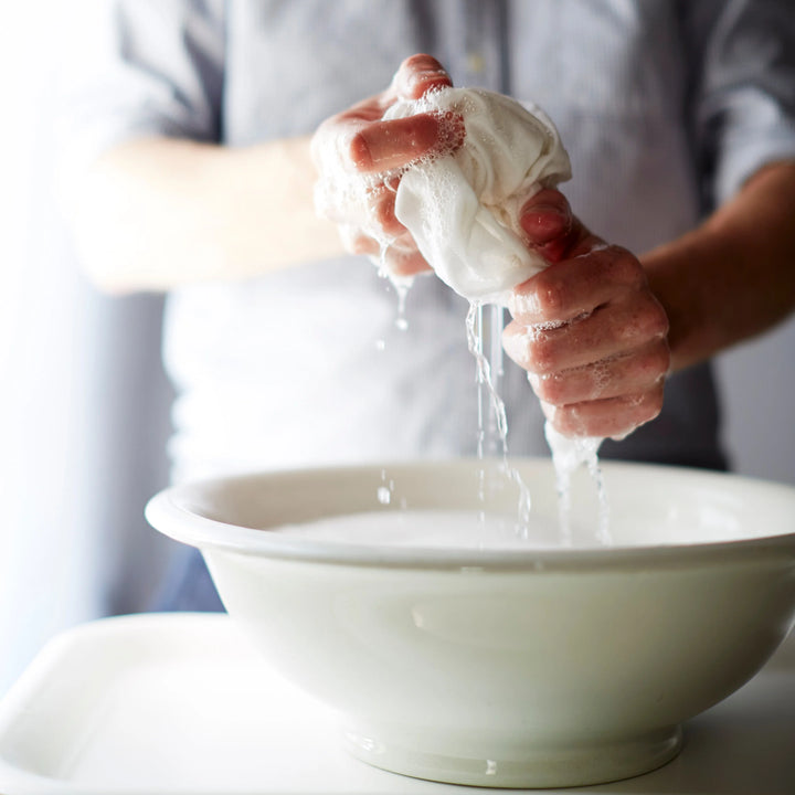 Someone with sensitive skin expertly squeezes a white cloth soaked in Common Goods unscented, plant-based 32 oz Laundry Detergent over a bowl, watching suds bubble up. They wear a light, long-sleeved shirt while handling the task skillfully.