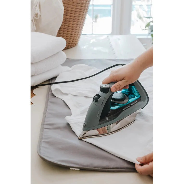 A person uses the Iron & Steam Blanket by Full Circle Home to iron a white shirt with a gray and teal steam iron. Folded white towels and a woven basket sit in the background, enhanced by bright natural light filtering through the window, creating a serene scene.