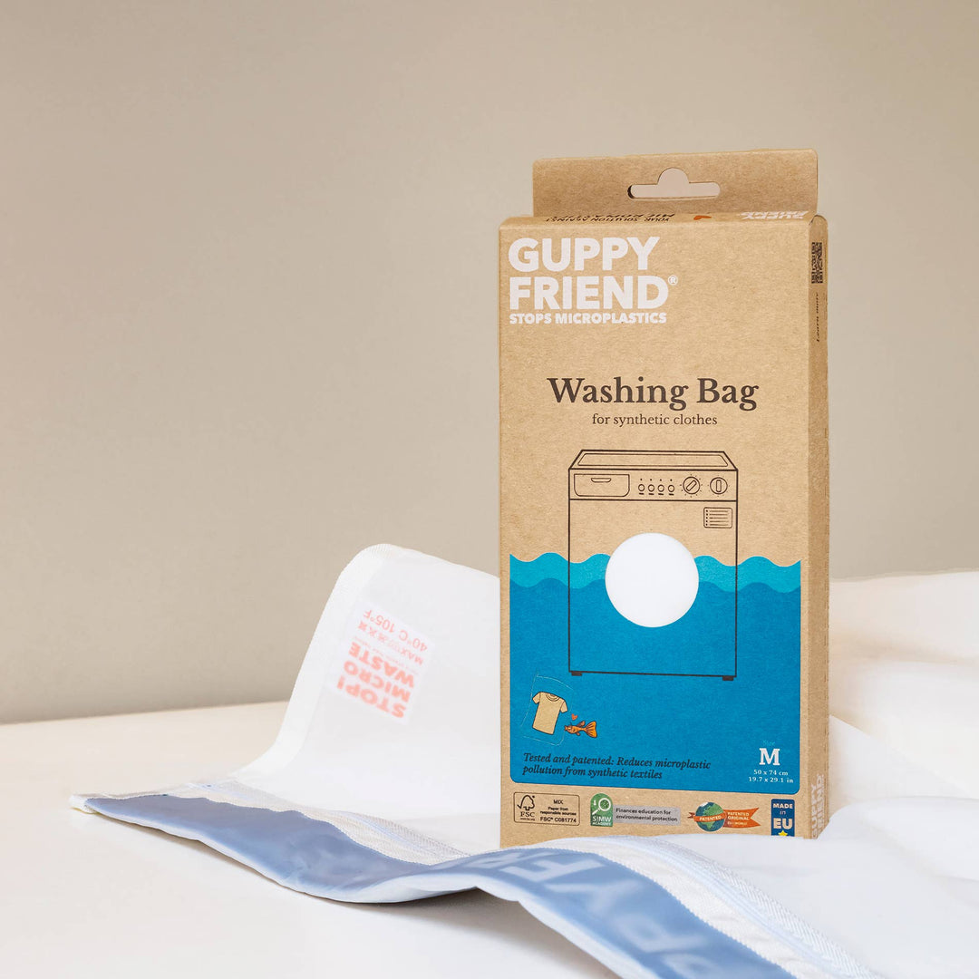 The image shows an upright cardboard box labeled GUPPYFRIEND Washing Bag-Medium 19.7 in. x 29.1 in. by GUPPYFRIEND, designed to combat microplastics from synthetic clothes, with a partially visible washing bag beside it on a neutral background, highlighting its fiber shedding reduction feature.