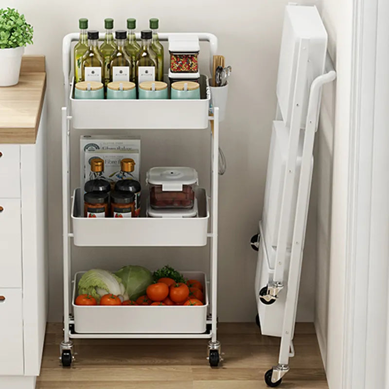 The All Things Laundry 3 Tier Folding Laundry Room Storage Cart with Handle and Roller Wheels in white holds wine bottles, cups, a spice jar, condiments, a plastic container, vegetables, and cabbage. A folded table leans against the wall beside this versatile cart.