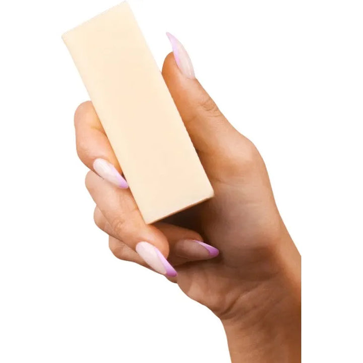 A hand with long, light pink nails holds a rectangular bar of off-white Vegan Laundry Stain Remover by Kind Laundry against a white background.