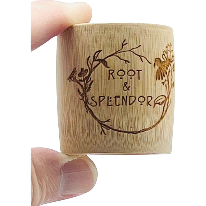 A hand cradles a small bamboo measuring cup, sustainably crafted, with Root and Splendor elegantly engraved. Intricate floral designs gracefully encircle the text, evoking its natural charm.