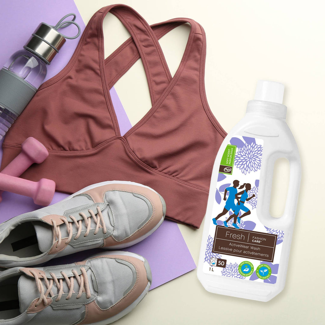 A pink sports bra, gray and pink sneakers, purple dumbbells, and a clear water bottle are arranged beside a 1L bottle of Fashion Cares Fresh® Activewear Wash, featuring eco-friendly ingredients and a floral label. All workout gear is neatly placed on a light surface, ready for action.