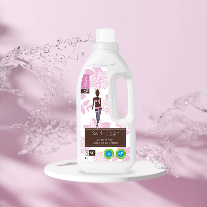 A bottle of Swirl Lingerie Wash - 1L by Fashion Care, featuring floral label designs, rests on a round white platform with water splashing against a pink backdrop. Made with eco-friendly ingredients, this detergent is ideal for lingerie care and lasts for up to 50 washes.